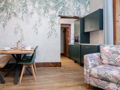 A dining area with a wooden table set with dishes and wine glasses next to green kitchen cabinets and a patterned sofa at Watermouth Castle West Tower Apartment in Berrynarbor near Ilfracombe