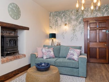 A living room with a green sofa pink and patterned cushions a woven coffee table with a blue bowl a wood stove and a wooden door at Watermouth Castle West Tower Apartment Berrynarbor near Ilfracombe