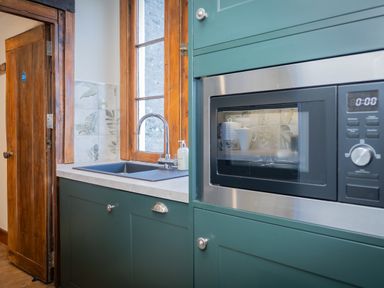 A kitchen area with green cabinets a built-in microwave a sink under a wooden framed window and a wooden door at Watermouth Castle West Tower Apartment Berrynarbor near Ilfracombe