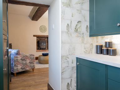 A kitchen with teal cabinets and floral patterned tiles next to a living room with a patterned armchair a round coffee table a brick fireplace and a wall clock at Watermouth Castle West Tower Apartment Berrynarbor near Ilfracombe