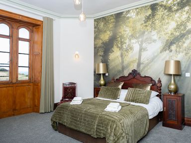 A bedroom with a wooden bed, green bedding, two lamps on wooden nightstands, a chair, and a large window at Watermouth Castle, West Tower Apartment in Berrynarbor near Ilfracombe