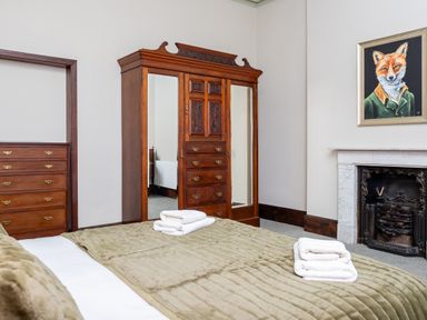 A bedroom with a bed covered by a beige quilt, wooden wardrobe with mirrored doors, fireplace, and a painting of a fox at Watermouth Castle West Tower Apartment Berrynarbor near Ilfracombe