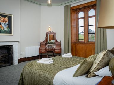 A bedroom with a bed covered in green bedding a wooden vanity desk with a chair a painting of a fox and a window with green curtains at Watermouth Castle West Tower Apartment Berrynarbor near Ilfracombe