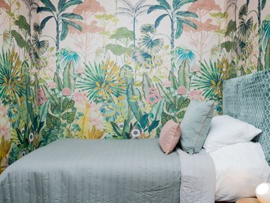A bedroom with a bed covered in a light blue quilt and pillows against a wall with tropical leaf wallpaper at Watermouth Castle West Tower Apartment in Berrynarbor near Ilfracombe