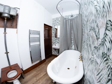 A bathroom with a clawfoot bathtub and shower curtain a wooden toilet seat a towel warmer and tropical leaf patterned wallpaper at Watermouth Castle West Tower Apartment in Berrynarbor near Ilfracombe