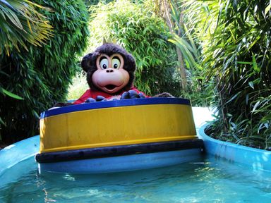 A person in a monkey costume riding in a yellow and blue boat on a water ride surrounded by green foliage