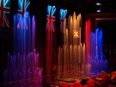 A lighted indoor water fountain show with multiple colored jets and British flags hanging in the background