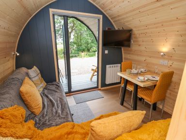 An interior of a wooden pod with a gray sofa and orange cushions a small dining table with two orange chairs and a wall-mounted television at Heron Pod in Nantmawr near Trefonen