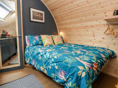 A bedroom with a double bed with floral bedding wood paneling walls a small shelf with hangers and a door leading to a kitchen area at Heron Pod in Nantmawr near Trefonen
