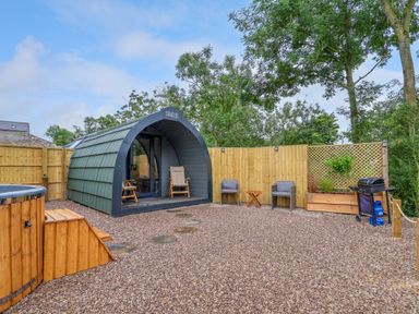 A small pod cabin with a wooden hot tub and outdoor seating area with grill in a fenced garden at Heron Pod in Nantmawr near Trefonen
