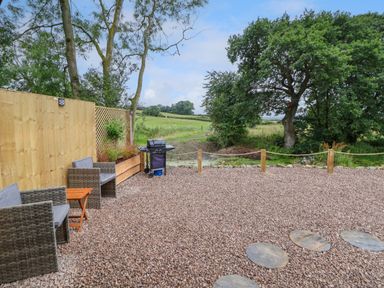 An outdoor patio area with wicker chairs a small wooden table a barbecue and trees beyond a wooden fence at Heron Pod in Nantmawr near Trefonen