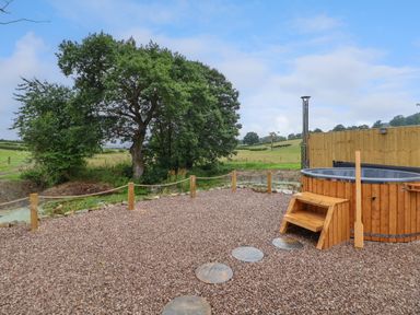 An outdoor gravel area with a wooden hot tub and steps near trees and fields at Heron Pod in Nantmawr near Trefonen