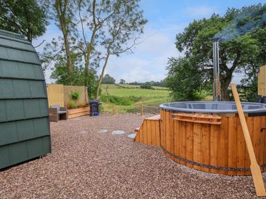 An outdoor area with a wooden hot tub a barbecue grill chairs and a small green cabin at Heron Pod in Nantmawr near Trefonen