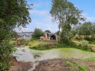 A small pond with green algae surrounded by grass and trees with a wooden pod and buildings in the background at Heron Pod in Nantmawr near Trefonen