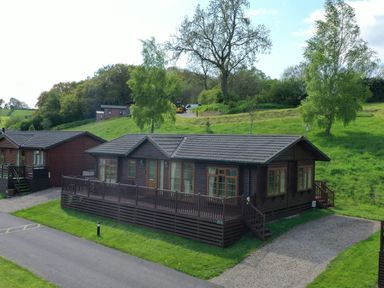 A wooden cabin with a deck and a gravel driveway surrounded by green grass and trees at The Fairhaven in Tunstall near Catterick North Yorkshire