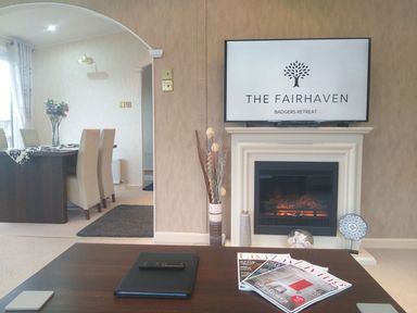 A living room with a fireplace and a television showing The Fairhaven Badgers Retreat above it and a dining area with chairs and a table in the background at The Fairhaven in Tunstall near Catterick North Yorkshire