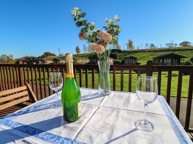 A table with a bottle, two wine glasses, and a vase with flowers on a balcony overlooking grassy hills and wooden cabins at The Fairhaven in Tunstall near Catterick, North Yorkshire