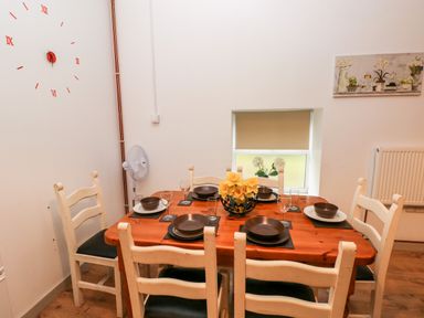 A dining room with a wooden table set for six with plates bowls glasses a flower centerpiece a wall clock and a window at Barn 2 Ystradgynlais