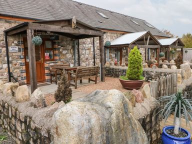 A stone cottage exterior with wooden porches and outdoor seating surrounded by a low stone wall and potted plants at Barn 3 Cwm-twrch Isaf near Ystradgynlais