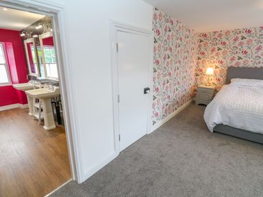 A bedroom with floral wallpaper a bed a nightstand with a lamp and an adjacent bathroom with two sinks at The Old Post Office in Praze-An-Beeble
