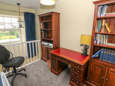 A study area with a wooden desk, bookshelves with books, a black office chair, a window with blue curtains, and a lamp at The Old Post Office in Praze-An-Beeble