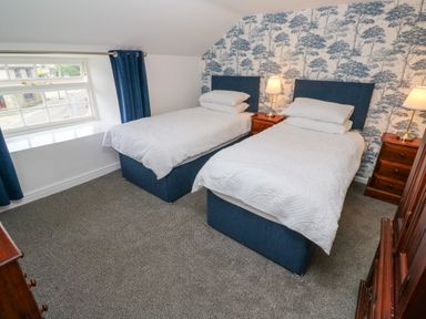 A bedroom with two single beds bedside tables lamps a window with blue curtains and patterned wallpaper at The Old Post Office in Praze-An-Beeble