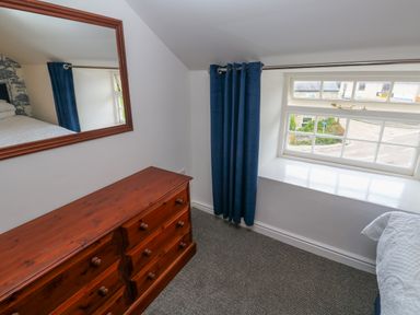 A bedroom corner with a wooden dresser a wall mirror blue curtains and a window at The Old Post Office in Praze-An-Beeble