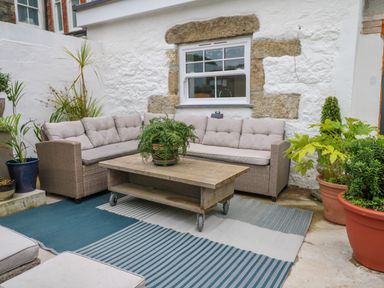 An outdoor seating area with a cushioned wicker sofa a wooden coffee table on wheels and several potted plants at The Old Post Office in Praze-An-Beeble