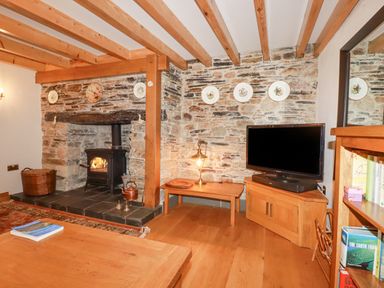 A living room with a stone fireplace wood stove wooden beams a television on a corner cabinet and a bookshelf at Mill Cottage in St Neot