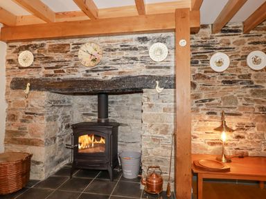 A stone fireplace with a wood burning stove a basket a metal bucket a kettle and a wooden table with a lamp and plates on the wall at Mill Cottage in St Neot