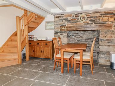 A dining area with a wooden table and four chairs next to a stone fireplace and wooden staircase at Mill Cottage in St Neot