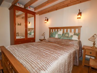 A bedroom with a wooden bed frame, side tables with lamps, a wooden wardrobe with mirrored doors and ceiling beams at Mill Cottage in St Neot