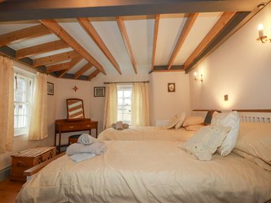 A bedroom with two beds, a dressing table, curtains on windows, and exposed wooden ceiling beams at Mill Cottage in St Neot