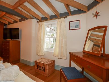 A room with wooden beams on the ceiling a wooden dresser with a television a window with curtains a wicker chest and a wooden dressing table with a mirror and a stool at Mill Cottage in St Neot