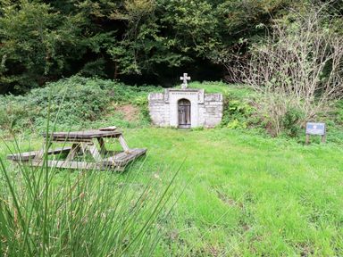 A stone structure with a cross and a wooden door surrounded by greenery with a wooden picnic table in front at Mill Cottage in St Neot