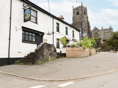 A stone building with a pub sign and wooden planter boxes outside with a stone church in the background at Mill Cottage in St Neot