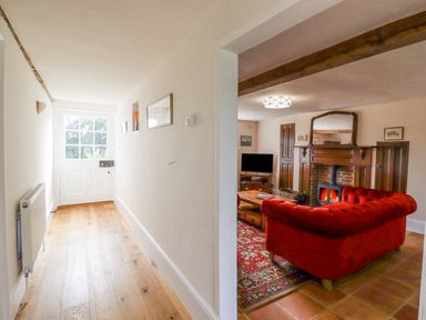 A hallway with wooden flooring leads to a white door and opens into a living room with a red sofa a wooden coffee table a fireplace and a television at West House Farm in Leiston