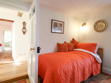 A small bedroom with an orange quilt and pillows on a single bed and a door open to a hallway at West House Farm in Leiston