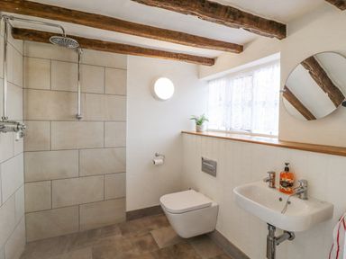 A bathroom with a wall-mounted toilet a small sink a round mirror a window with lace curtains and exposed wooden ceiling beams at West House Farm in Leiston