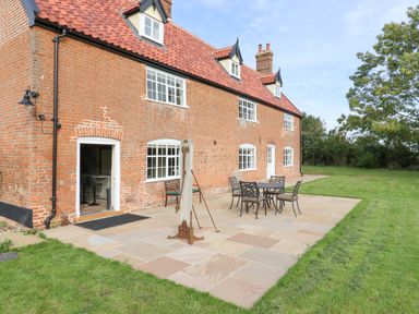 A brick house with a tiled roof and white framed windows a patio with a metal table and chairs and a grassy lawn at West House Farm in Leiston