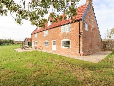 A brick house with a red tiled roof and outdoor furniture on a patio in a grassy yard at West House Farm in Leiston