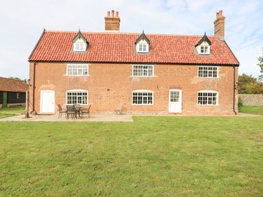 A brick house with red roof tiles and white framed windows with outdoor furniture on a patio and a green lawn at West House Farm in Leiston