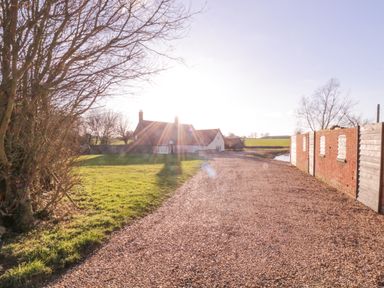 A gravel driveway leading to a house with a brick wall and wooden fence on the right and grass and trees on the left at West House Farm in Leiston