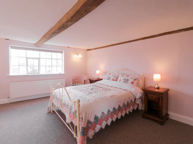 A bedroom with a double bed with patterned bedspread and pillows two wooden bedside tables with lamps a pink armchair beneath a window with wooden beams on the ceiling at West House Farm Leiston