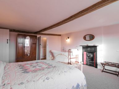 A bedroom with a bed covered with a quilt, a wooden wardrobe with a mirror, a white bathtub, a black fireplace, and exposed ceiling beams at West House Farm in Leiston