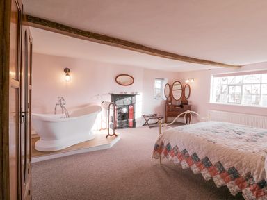 A bedroom with a freestanding bathtub a wooden dresser with mirrors a bed with a quilt and a fireplace at West House Farm in Leiston