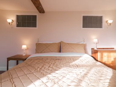 A bedroom with a bed covered with beige bedding two nightstands with lamps wooden beam on the ceiling and two small windows at West House Farm in Leiston