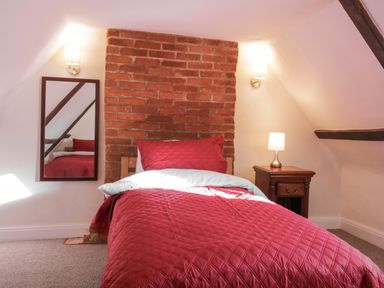 A bedroom with a single bed with red bedding a wooden nightstand with a lamp a brick wall and a mirror at West House Farm Leiston
