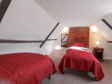A bedroom with two single beds with red bedspreads a wooden nightstand with a lamp a wall mirror and exposed brick at West House Farm in Leiston