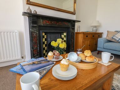 A living room with a wooden coffee table holding cupcakes, scones, mugs, plates and books in front of a fireplace and a blue sofa at Blue Yonder in Weymouth
